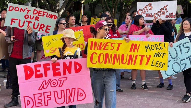 Several people gathered outside with colourful signs with messages like "defend not defund" and "invest in community."