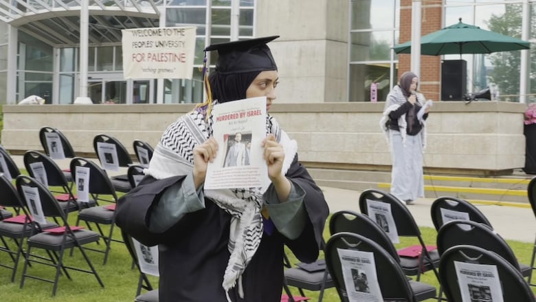 A woman wear a commencement cap and gown while holding a poster that says a person's name and the caption 'Murdered by Isreal'