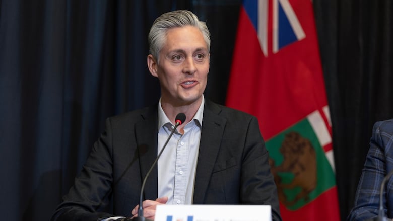 a man with grey hair wearing a black suit speaks into a microphone in a conference hall with a flag behind him