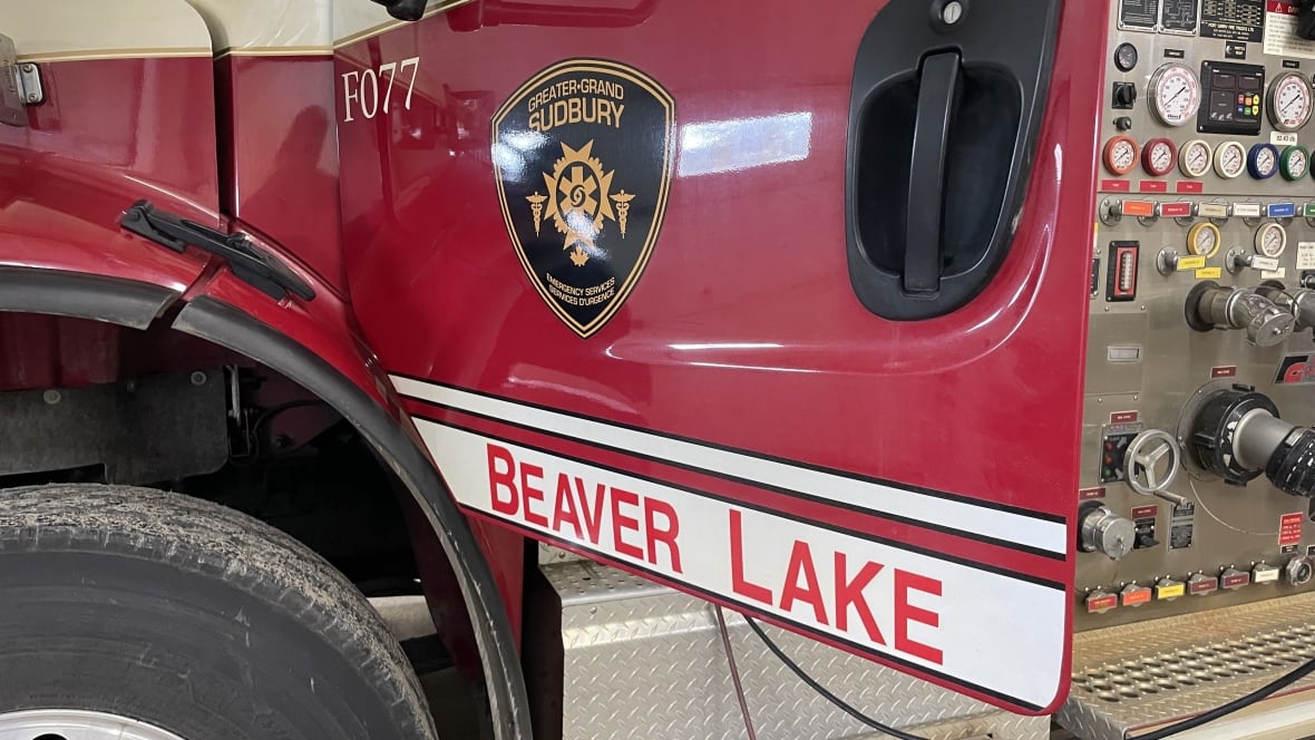 A side view of a red fire truck with the door bearing the black and gold logo of the Sudbury fire service and a banner saying Beaver Lake.