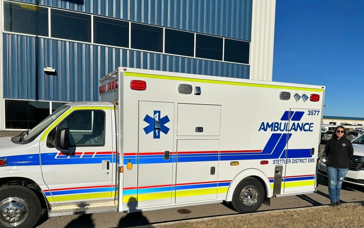 A white ambulance with blue writing parked outside on a sunny day with a woman standing next to it.