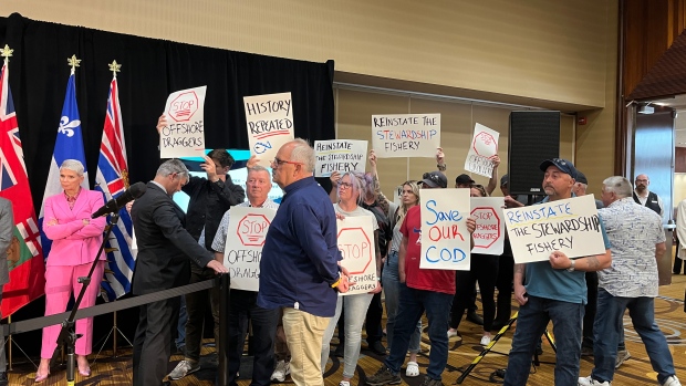 Newfoundland fish harvesters crash news conference to demand reinstating of cod moratorium; A group of about 15 people stood next to the ministers with signs calling for the stewardship fishery to be reinstated\. FFAW President Greg Pretty, seen in the front wearing a blue shirt, said harvesters can't let Ottawa's decision stand\.