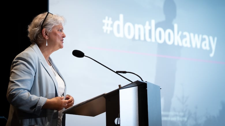 A woman stands at a podium in front of a screen that says "don't look away."