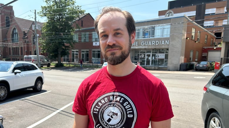 A man stands in front of a city street. He wears a red shirt and has a beard. he is balding . 