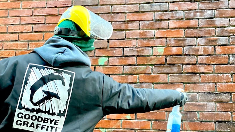 Person wearing a jacket with the words "Goodbye Graffiti" uses a spray bottle to clean graffiti on a brick wall.