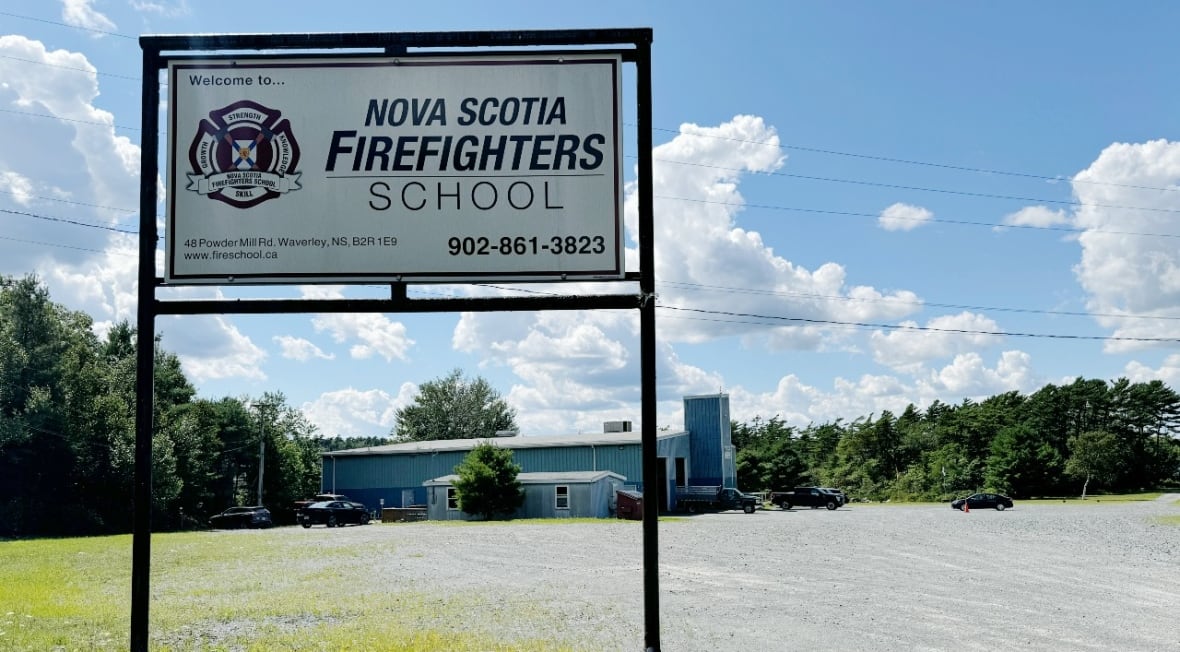 A white sign stands against a blue sky with the school's crest and name on it. A large blue industrial building is in the background