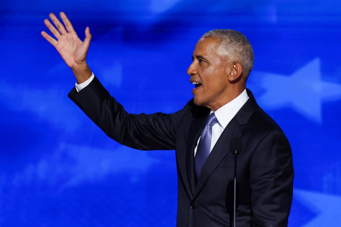 A man with grey hair waves and smiles at a podium.