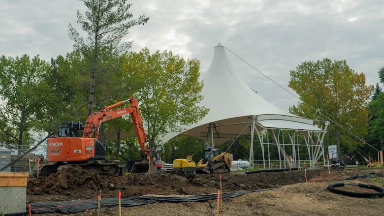 A view of a white tent outdoors under a cloudy sky, with the ground dug up in front of it and an orange excavator.