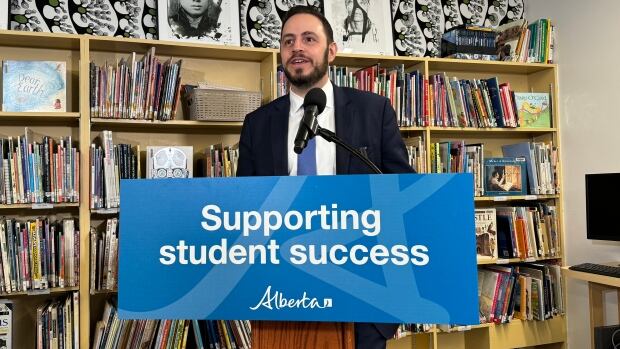 Education Minister Demetrios Nicolaides will announce on Saturday that 11 new school construction projects are moving ahead. Five of the projects are in Edmonton, three are in Calgary, and three are in Calgary bedroom communities. A man with dark hair and dark facial hair wearing a dark-coloured suit, white shirt, and blue tie stands behind a podium that reads "supporting student success." Behind him are brown wooden shelves filled with books.