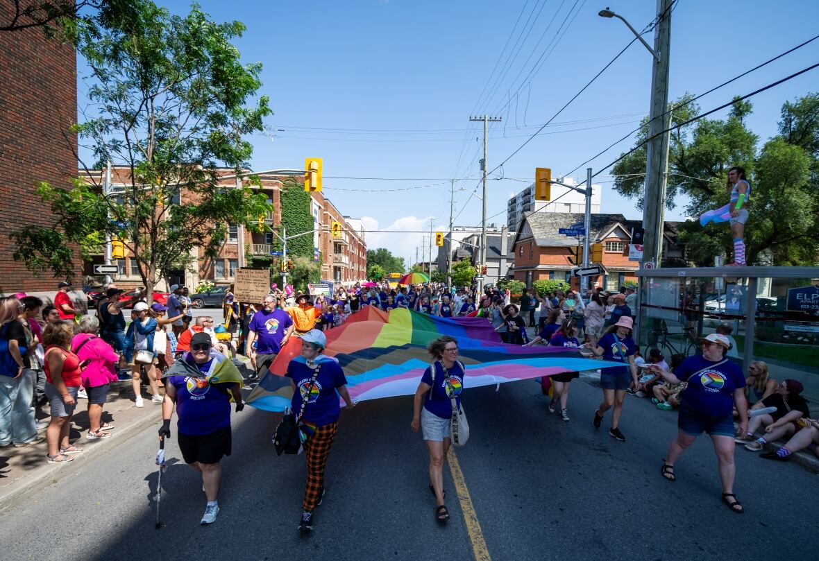 A crowd of people carry a large rainbow-coloured flag through a downtown street during a parade.