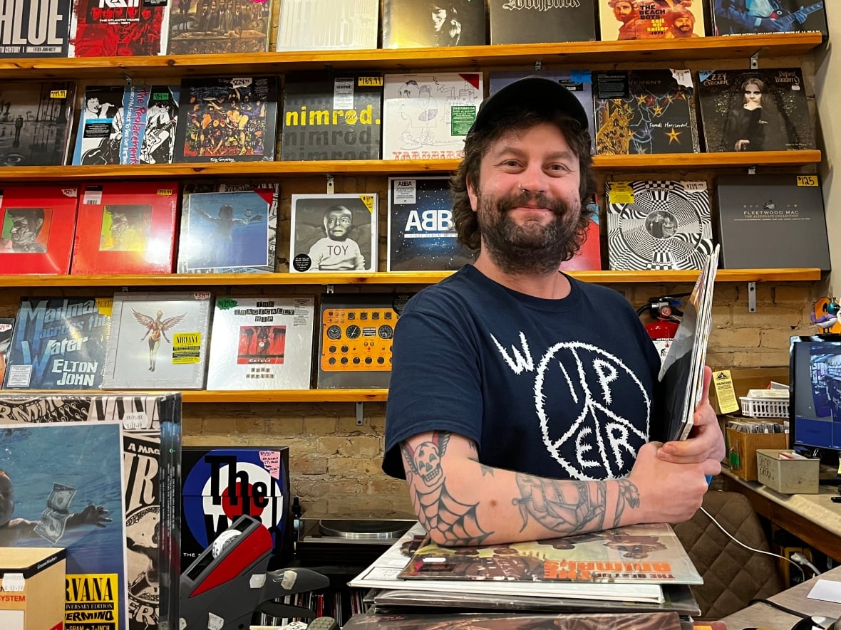 A man stands at the front counter of a store with records on the wall behind him. 