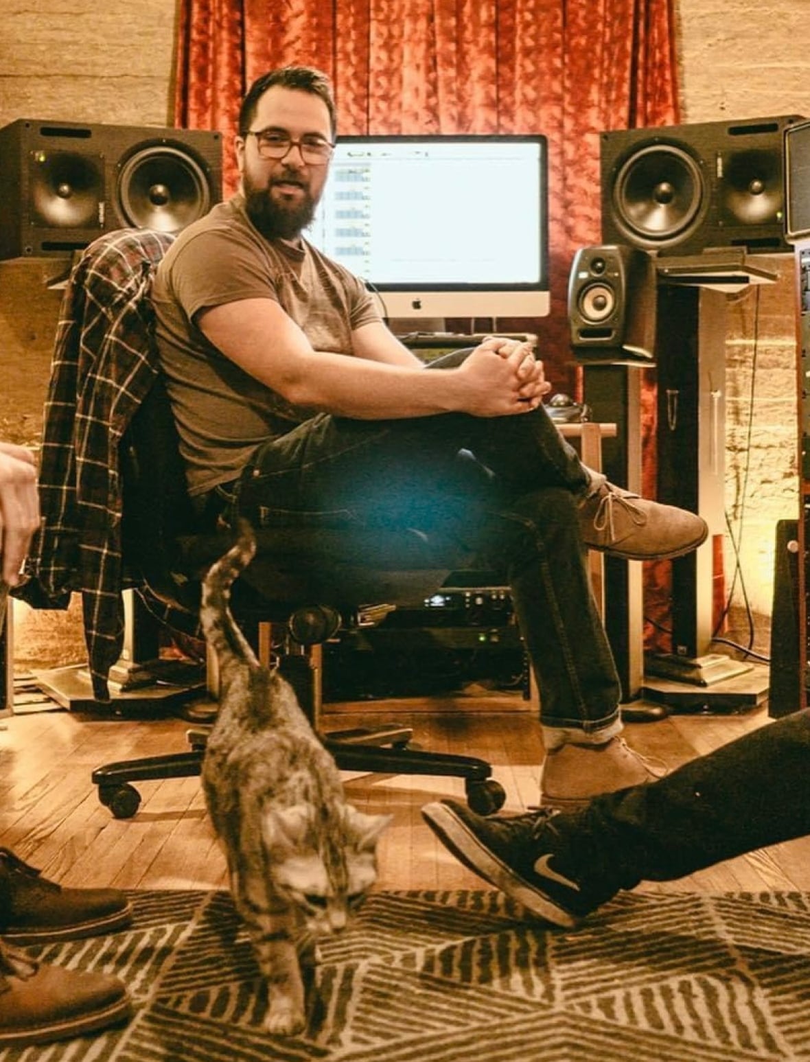 A man sits in a chair in a recording studio. There's a cat on the ground. 