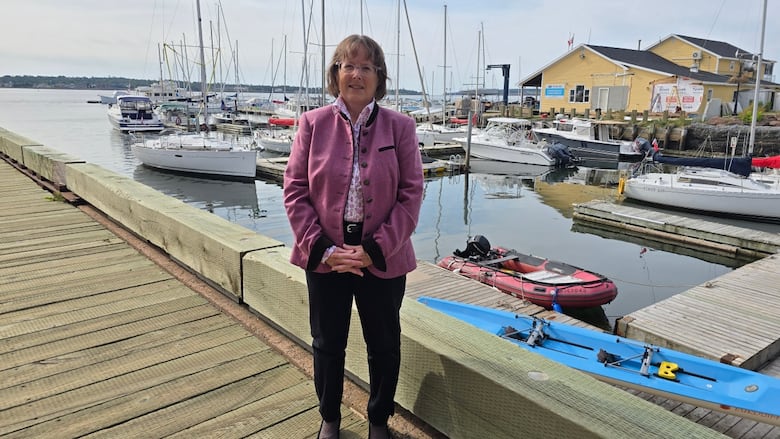 Woman with glasses and short brown hair in a pink jacket, standing on a wharf in front of sailboats.