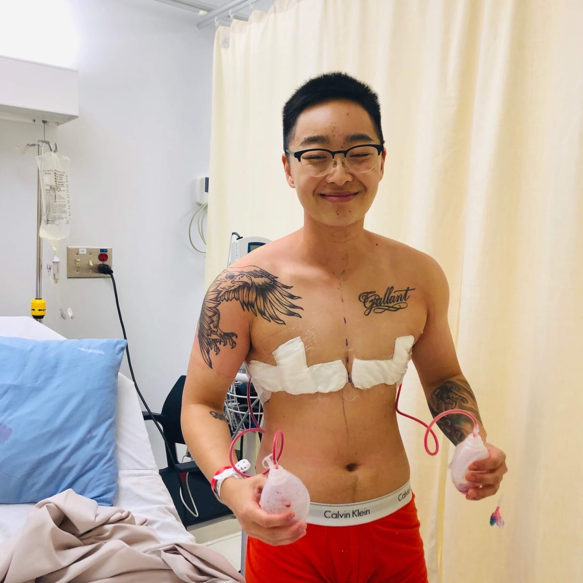 A man with bandages on his chest smiles as he stands in a hospital room.