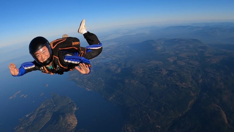 A man wearing a helmet and a blue and black suit smiles as he skydives over a mountainous coastline.