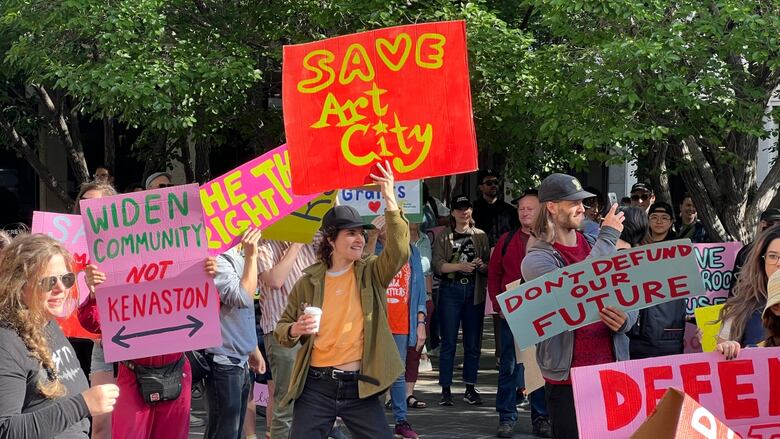 A crowd of people are standing in a court yard holding signs. One reads "Save Art City."