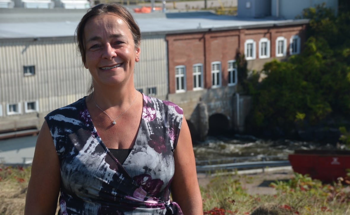 A smiling woman in a fashionable top stands in front of a hydroelectric dam 