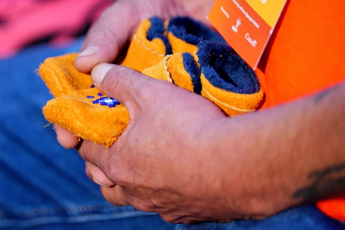 Someone holds a small pair of moccasins on their knees. Moccasins have a blue and white pearl pattern on them.