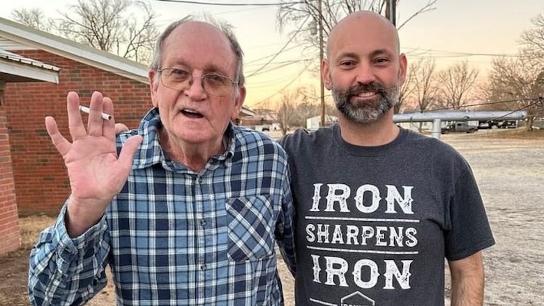 Two men, one elderly and one middle-aged, stand outside with their arms around each other's shoulders. The younger man is smiling and the older man is waving with a cigarette between his fingertips.
