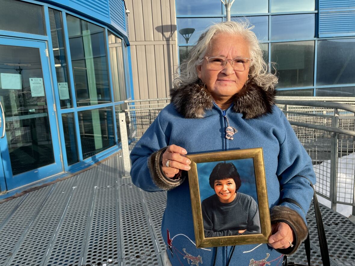 A woman wearing a parka with white hair holds a framed photograph of a young girl