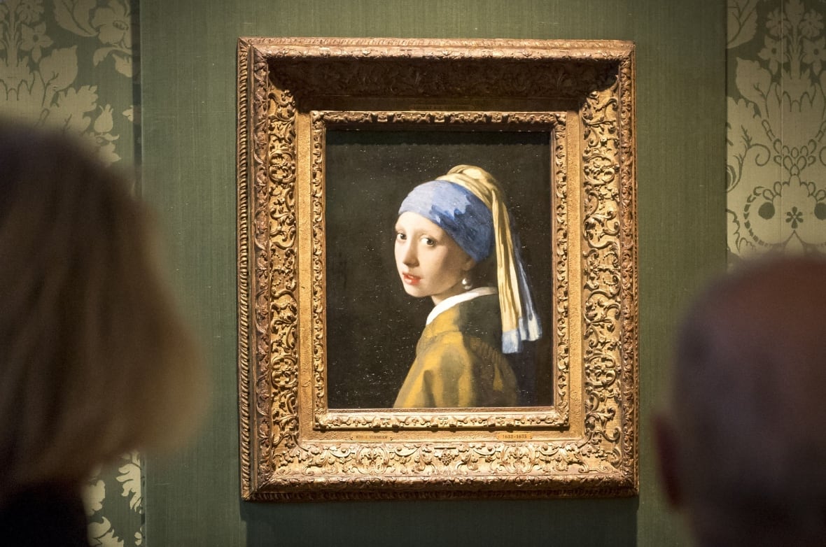The Girl with the Pearl Earring painting that shows a young girl against a black backdrop looking over her shoulder at you with large eyes and a single pearl earring visible that's as large as her eyes. The artwork is framed in an ornate gold frame, hanging on a green museum wall, with people in the foreground blurred out who are looking at the painting.