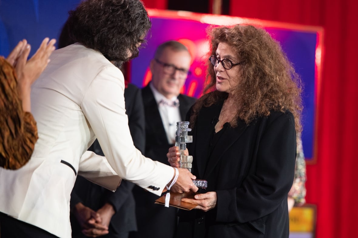 A white woman with curly hair and black glasses receives a glass trophy from a man with long black hair in a white tuxedo.