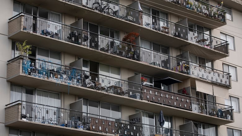 Multiple levels of a beige apartment building are pictured, filling the entire frame. Each level has a row of balconies, with glass sliding doors and an assortment of occupants' possessions.