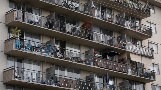 Multiple levels of a beige apartment building are pictured, filling the entire frame. Each level has a row of balconies, with glass sliding doors and an assortment of occupants' possessions.