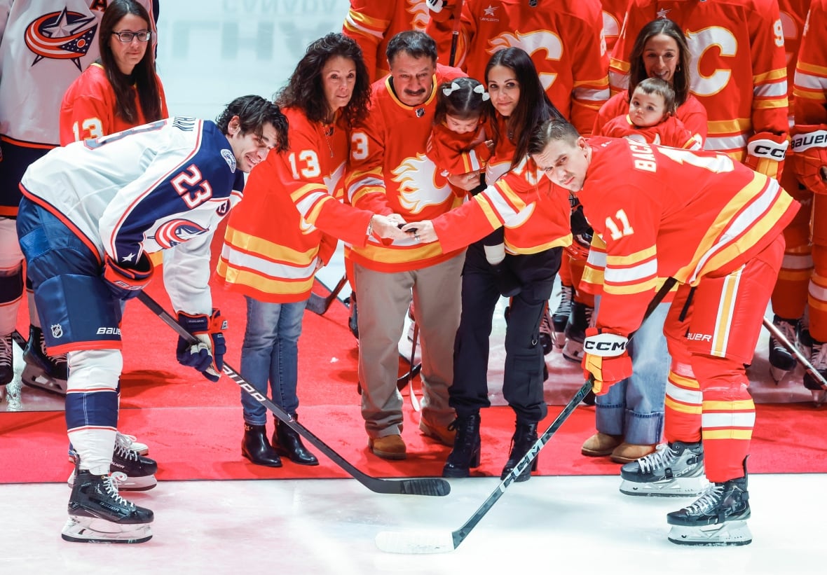 A group of people wearing red jerseys hold a puck over a faceoff between two men dressed in hockey equipment. One man wears white, the other wears red.