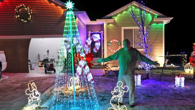 A smiling, elderly bald man stands in front of a house on a snow covered lawn with his arms outstretched, surrounded by Christmas lights and decorations, including two giant candy canes, a tree and a neon Mickey and Mini Mouse. 