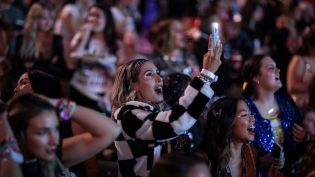 Fans, with the focus on girls in the foreground of the shot, cheer, while one holds up a cellphone toward the stage.