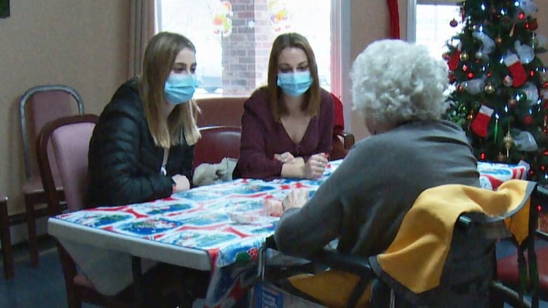 Two women wearing masks visit with a senior living in a long-term care home. The senior is in a wheelchair. There are Christmas decorations.