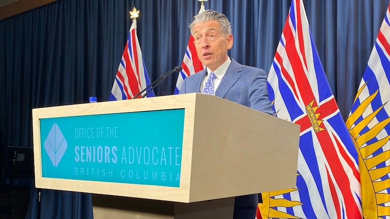 A man with grey hair speaks in front of a series of B.C. flags at a podium.