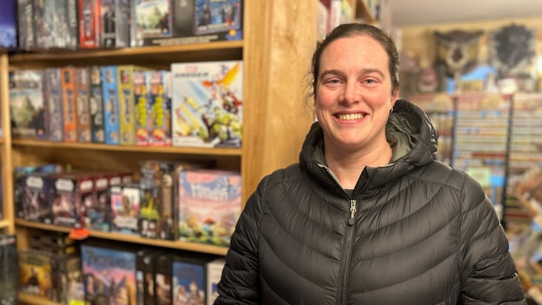 A woman with brown hair and a black jacket smiles at the camera, with toys and games on shelves behind her.