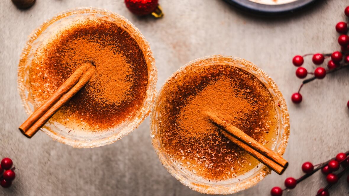 Top down shot of 2 glasses with vegan eggnog. They're dusted with nutmeg and rimmed with sugar, and each glass has a cinnamon stick in it. 