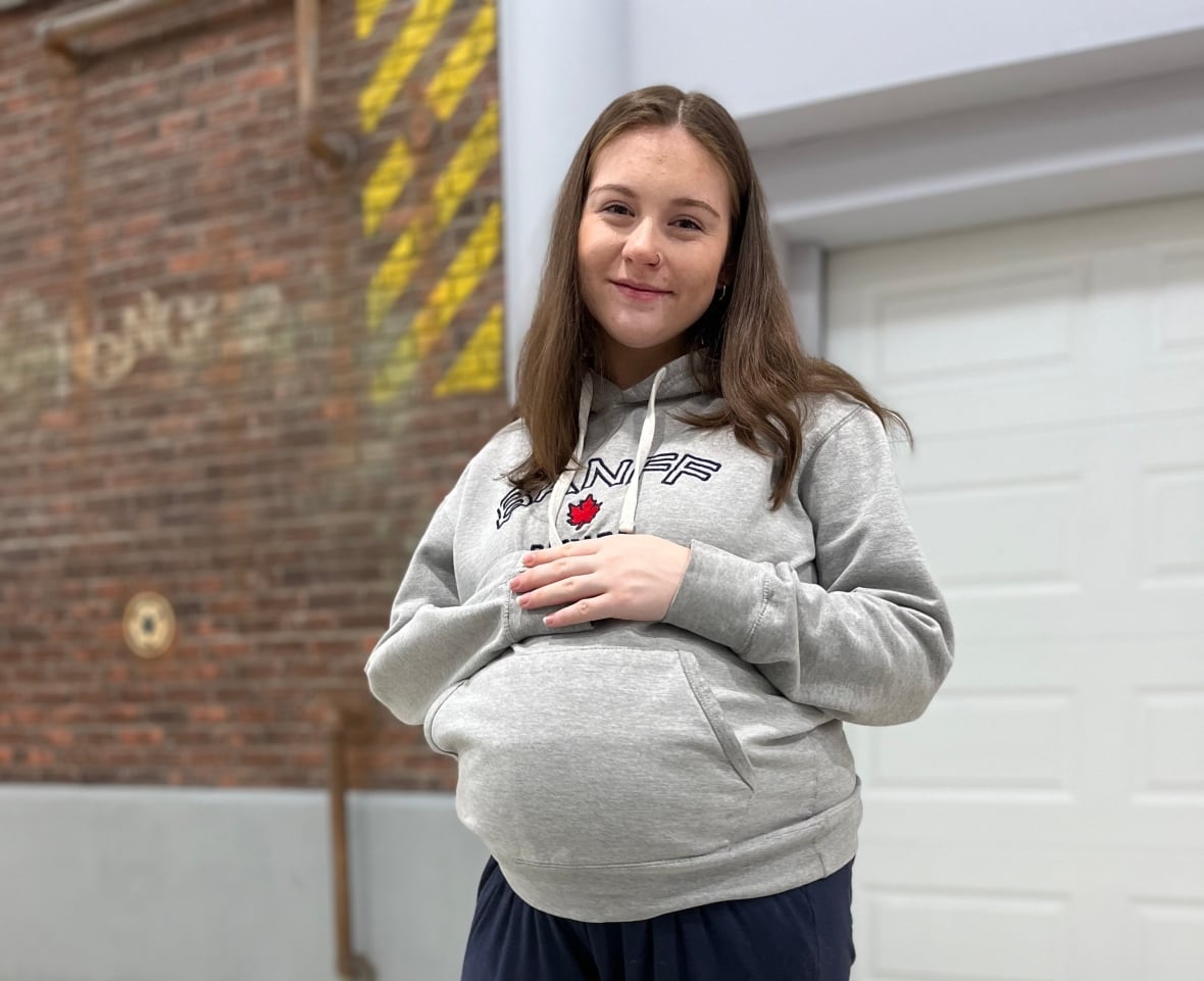 A smiling pregnant woman wearing a sweatshirt rests her folded arms at the top of her belly.