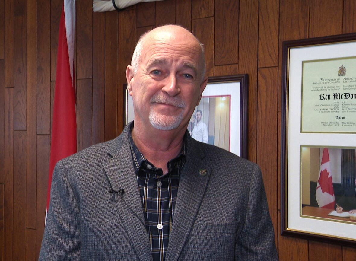 A smiling man wearing a plaid blazer stands in front of a wall with wood paneling. There are framed pictures on the wall and a Canadian flag behind his right shoulder.