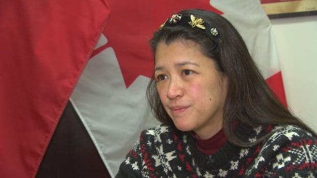 A woman in a knitted black, red and white sweater is seated at a table, in front of the Canadian and Manitoban flags.