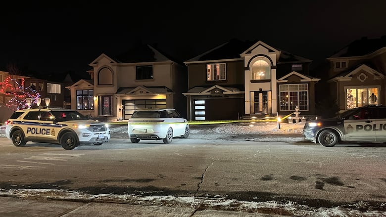 A suburban house at night with two police cars parked outside and a white car halfway pulled out of the driveway with police tape surrounding it.