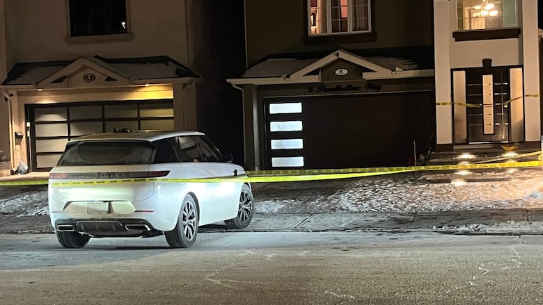 A suburban house at night with a white car halfway pulled out of the driveway with police tape surrounding it.