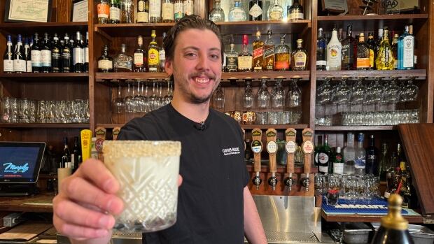 Caleb MacKinnon, manager of Gahan House Charlottetown, holds up a mocktail called Caught in the Rain as part of the bar's Dry January menu. A bartender with a black T-shirt on and dark curly hair smiles to camera as he holds up a milky cocktail.