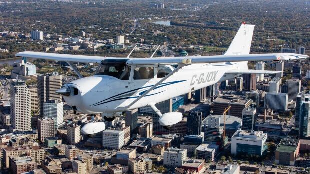 A small white aircraft marked C-GJOX on the side is seen flying over downtown Winnipeg above commercial and high-rise buildings, with trees and a river in view at a distance.