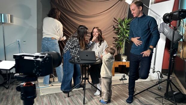 The volunteer crew for a local Calgary news show for Ukrainian newcomers get ready for their weekly shoot. From left to right, photo studio owner Luiza Yaromchuk steams the backdrop, make-up artist Nataliia Semenova does last minute touch-ups for host Anhelina Taran while Nataliia's three-year-old son Michaylo looks on and cameraman Anton Sholomytskyi fixes the lights. Five people get ready for a TV shoot, steaming the background curtains, fixing lights and doing make-up.
