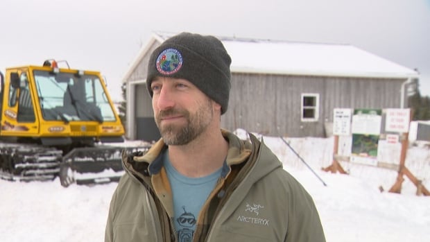 Chad Mooney wears a tuque and jacket. The trail grooming machine and an information board can be seen behind him.