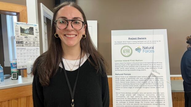 Tess Donohue, the Wejipek Wind Project manager, says A woman wearing glasses and a black sweater with a white t-shirt underneath stands with her arms behind her back.