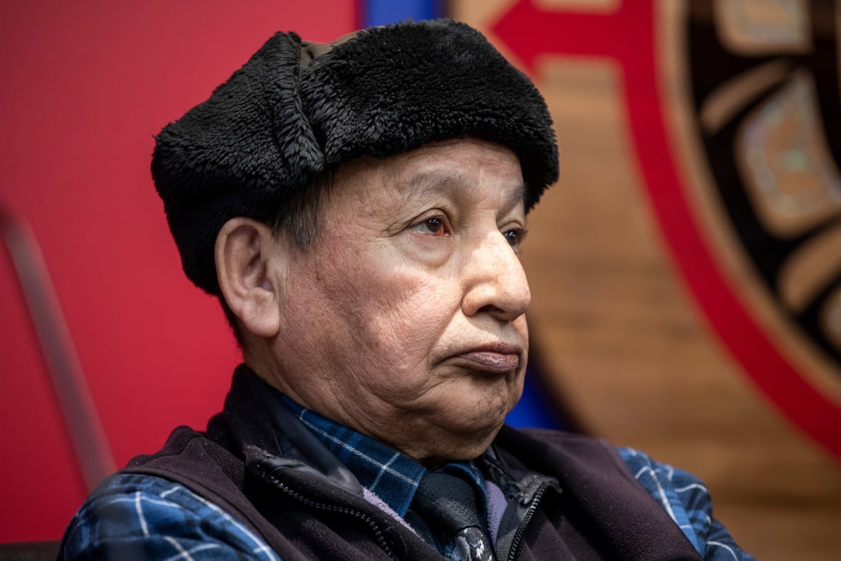 An Indigenous man wearing a black hat is pictured in profile, with a crest behind him.
