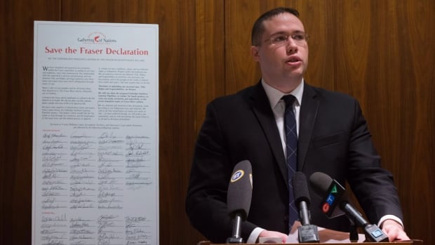 A man with glasses and a suit speaks at a podium next to a signed document.