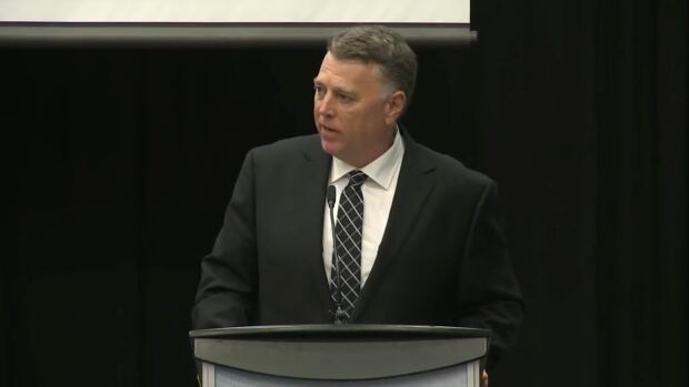 A man in a black suit and tie stands at a podium, addressing an audience