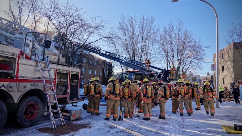 Dozens of firefighters mill near a firetruck with smoke billowing in the background. 