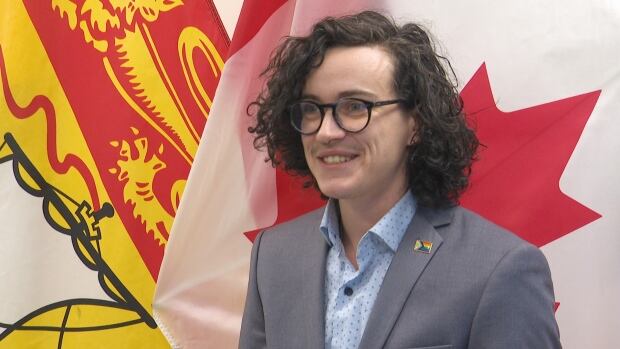 Mariah Darling, the education co-ordinator for Chroma New Brunswick, said they're 'proud that New Brunswick has taken a different path.' A person with black glasses and curly black hair wearing a grey suit jacket and baby blue shirt. They are standing in front of a New Brunswick flag, left, and a Canada flag, right.